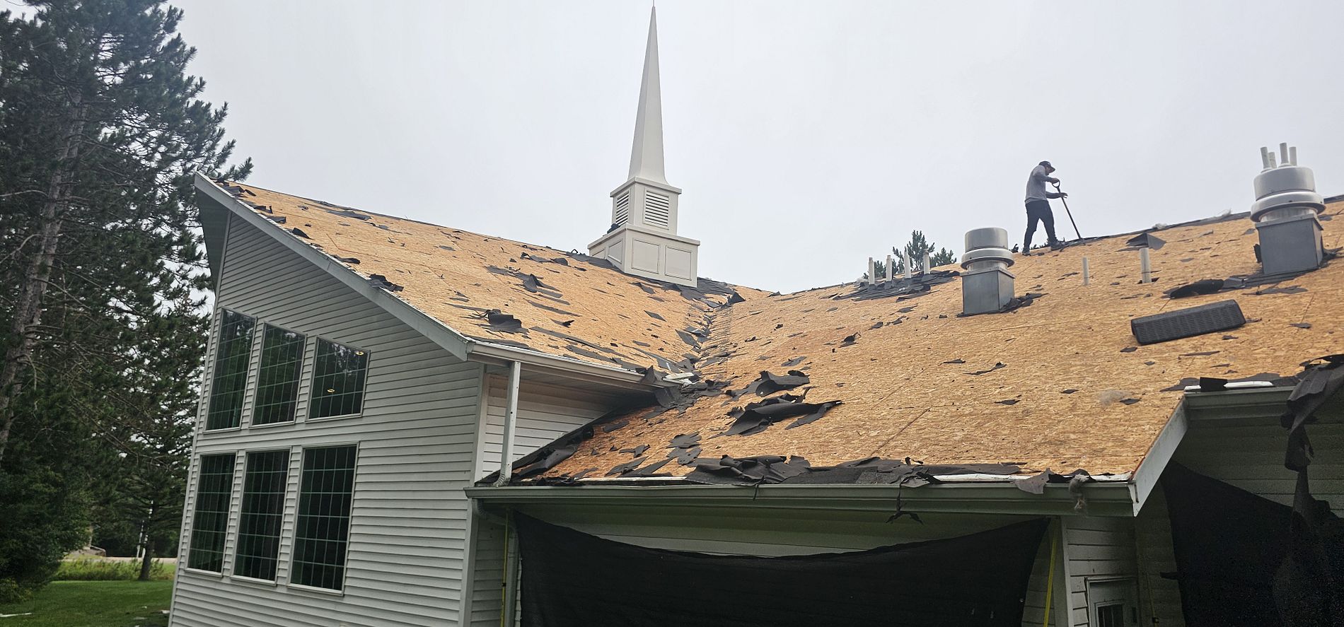 Large church roof in Merrill, WI during tear-off with wind-damaged shingles removed and roof decking exposed.