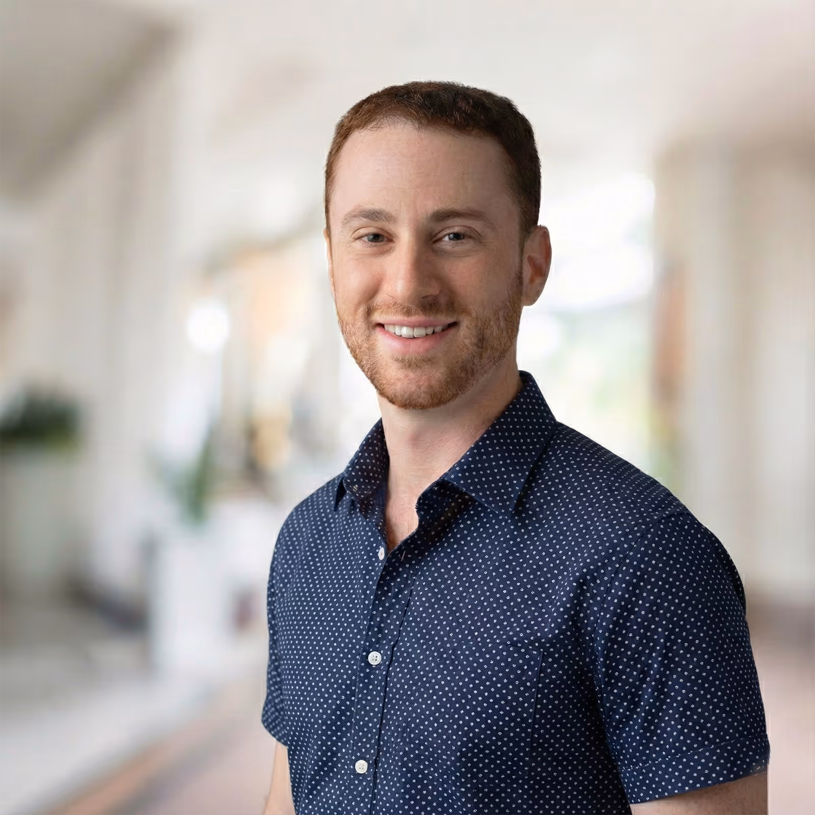 Smiling man with short red hair and beard wearing a navy blue shirt with white dots against a blurred indoor background.