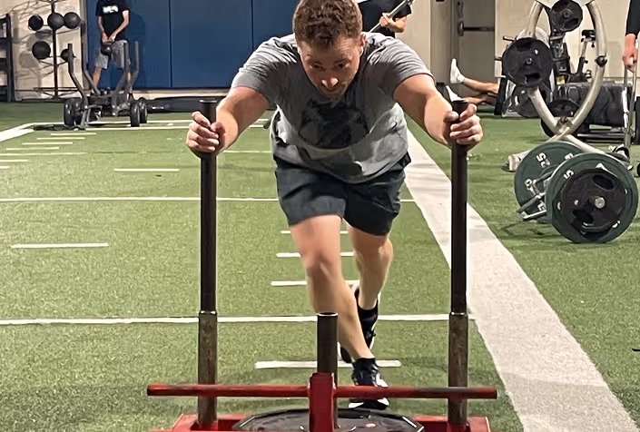 Man pushing a weighted sled forward on a green turf indoor gym floor.