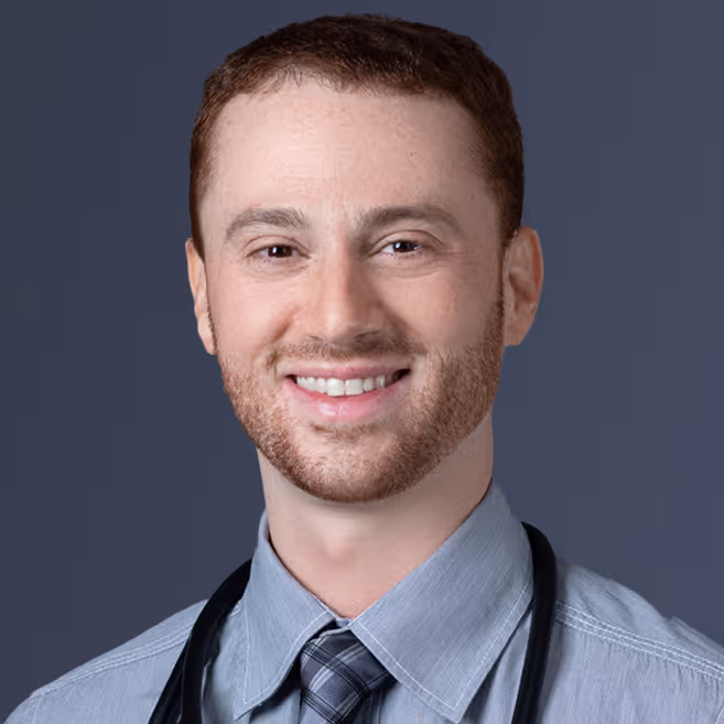 Smiling young man with short reddish hair and beard wearing a gray shirt, tie, and stethoscope.