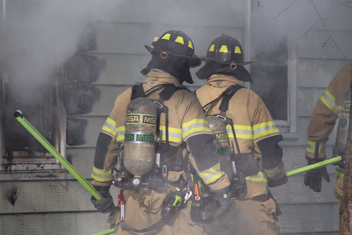 Group of firefighters in protective gear standing in front of a Burnsville Fire ladder truck on a wet concrete surface under a cloudy sky.
