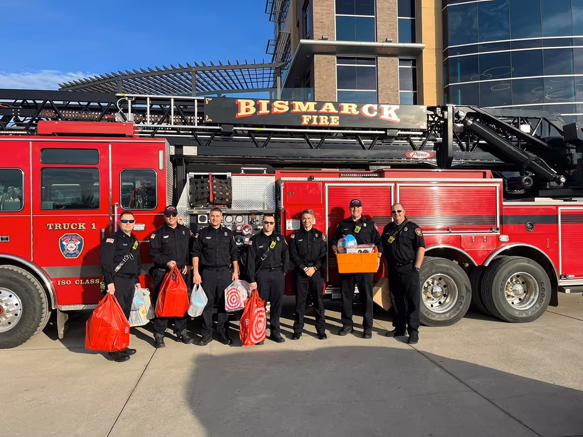 Seven firefighters in black uniforms standing in front of a red Bismarck fire truck holding bags and a box of supplies.