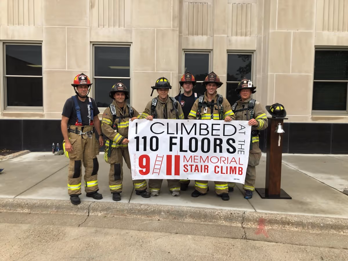 Six firefighters in full gear holding a banner that reads 'I climbed 110 floors at the 9/11 Memorial Stair Climb' outside a building.