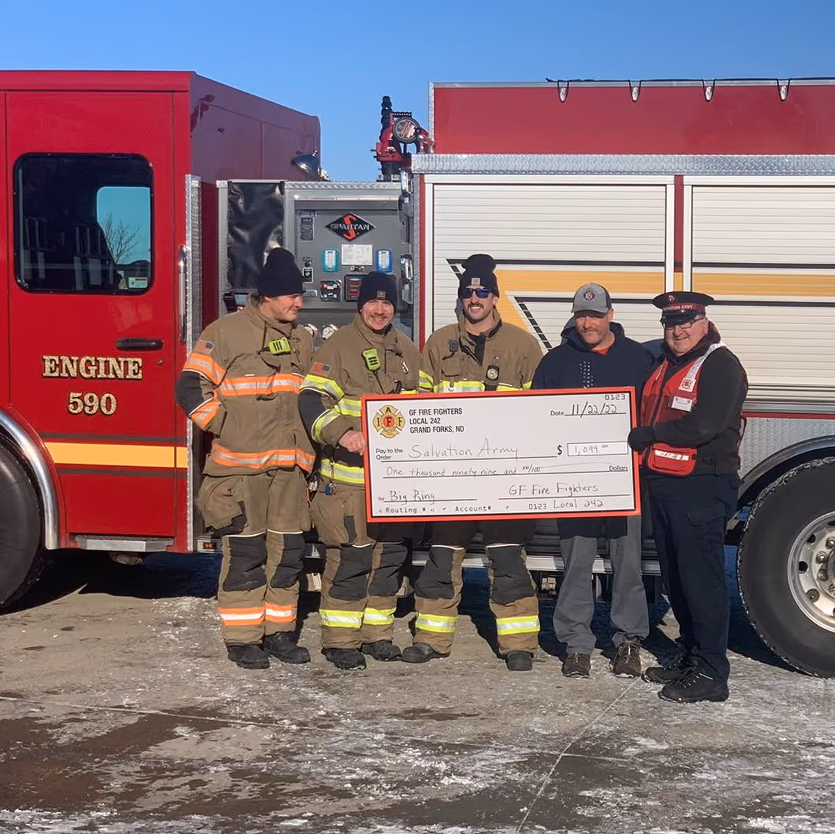 Four firefighters and one Salvation Army representative holding a large donation check in front of a red fire engine labeled Engine 590.