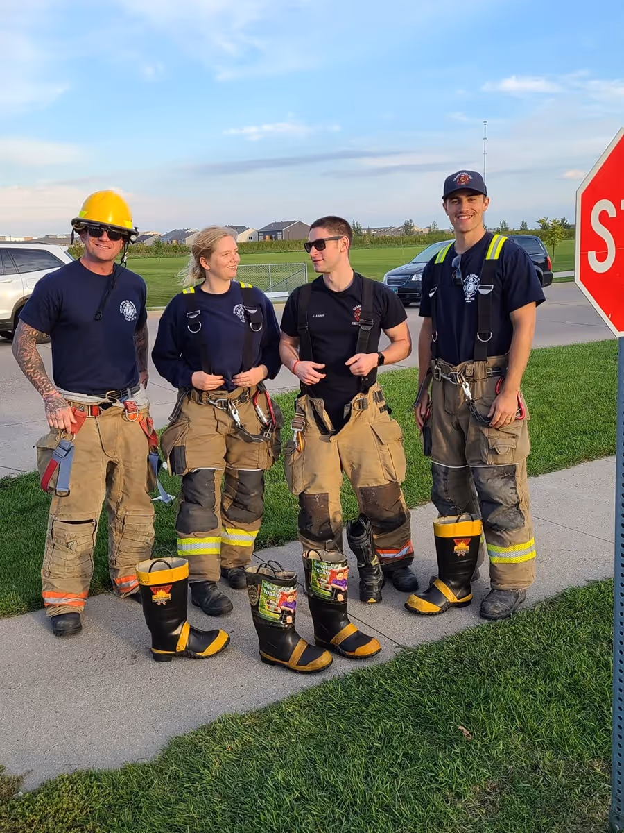 Four firefighters wearing navy shirts and tan protective pants stand smiling on a sidewalk next to fire boots lined up in front of them near a stop sign.