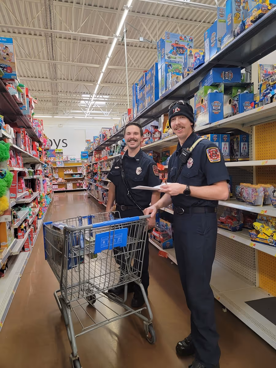 Two uniformed firefighters with a shopping cart standing in a toy aisle of a store.