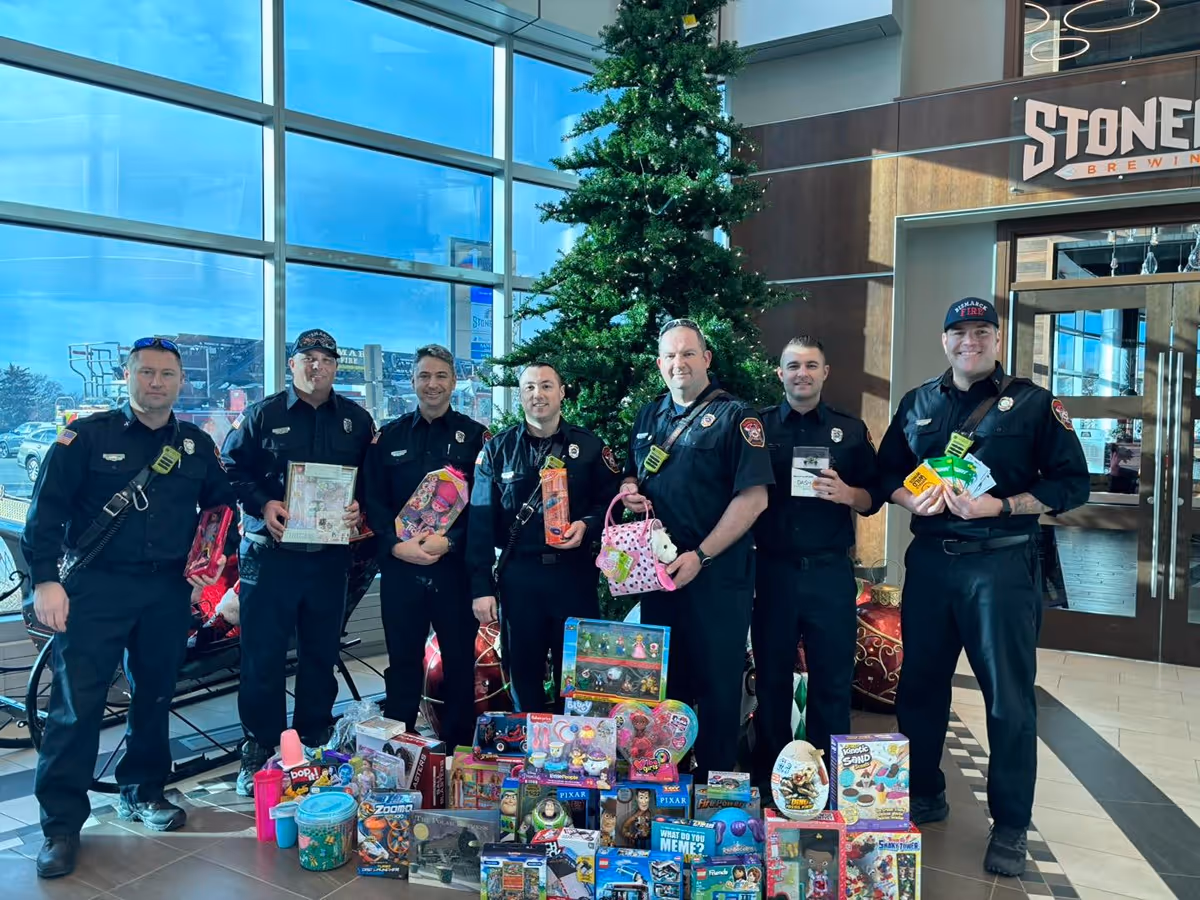 Seven firefighters in uniform standing indoors by a Christmas tree with a large collection of donated toys in front of them.