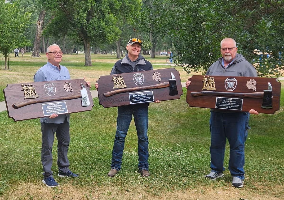 Three men standing outdoors on grass holding large wooden plaques with mounted axes and commemorative badges.