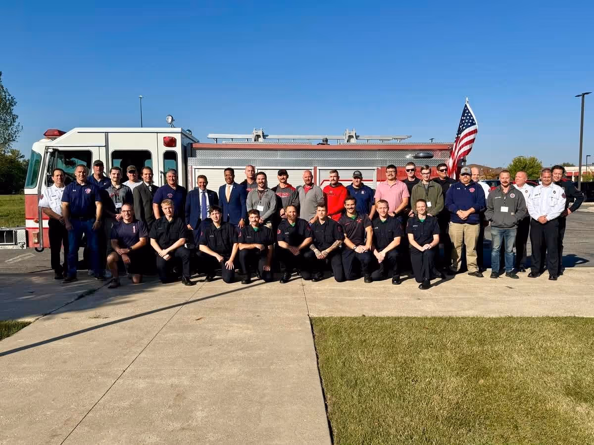 Group of firefighters and officials posing in front of a fire truck with an American flag on a sunny day.