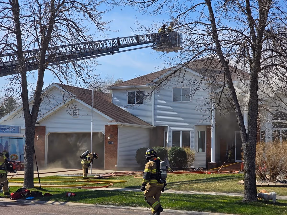 Firefighters responding to a house fire with smoke coming from the garage and front door, using a ladder truck and hoses.