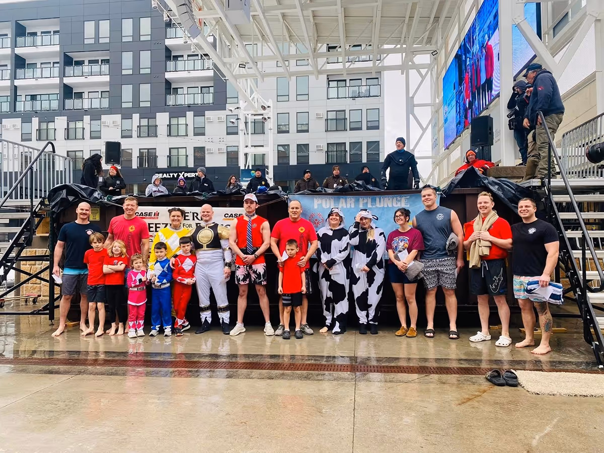 Group of people, including children and adults dressed in costumes such as Power Rangers and cow onesies, posing in front of a Polar Plunge event setup in an urban area.