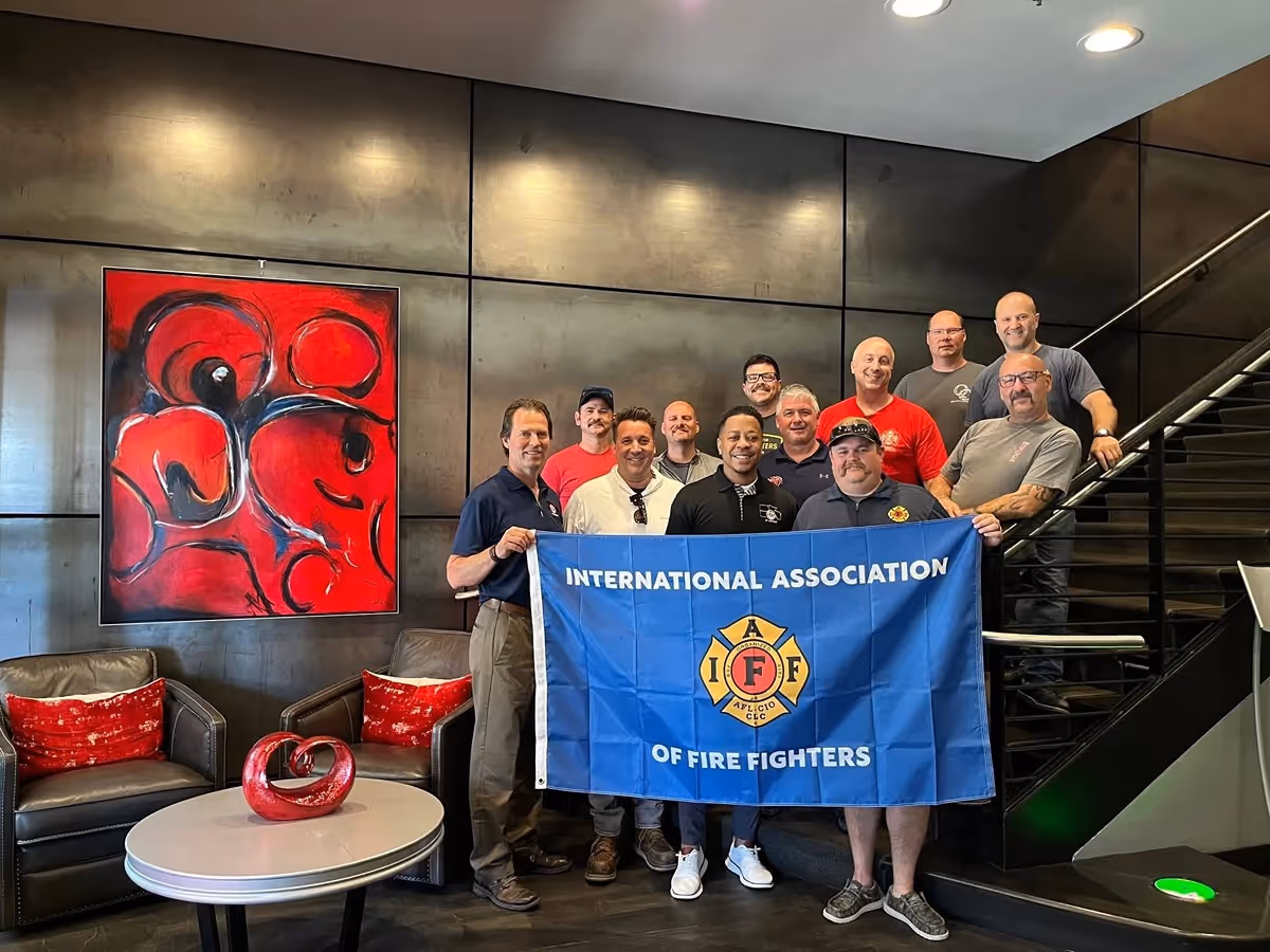 Group of men posing indoors holding an International Association of Fire Fighters flag, with modern art and furniture in the background.