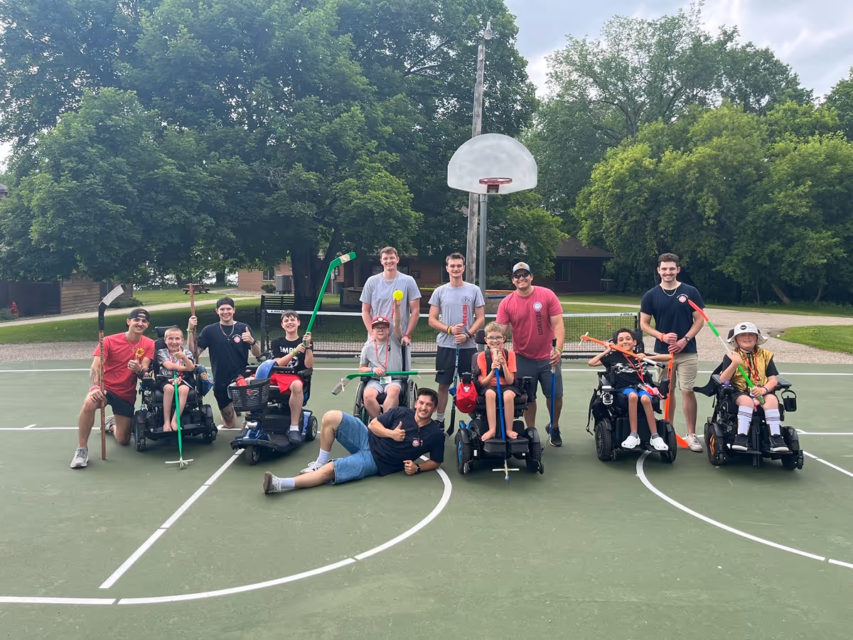 Group of children in wheelchairs and adults posing together on a basketball court holding floor hockey sticks, surrounded by trees and outdoor buildings.