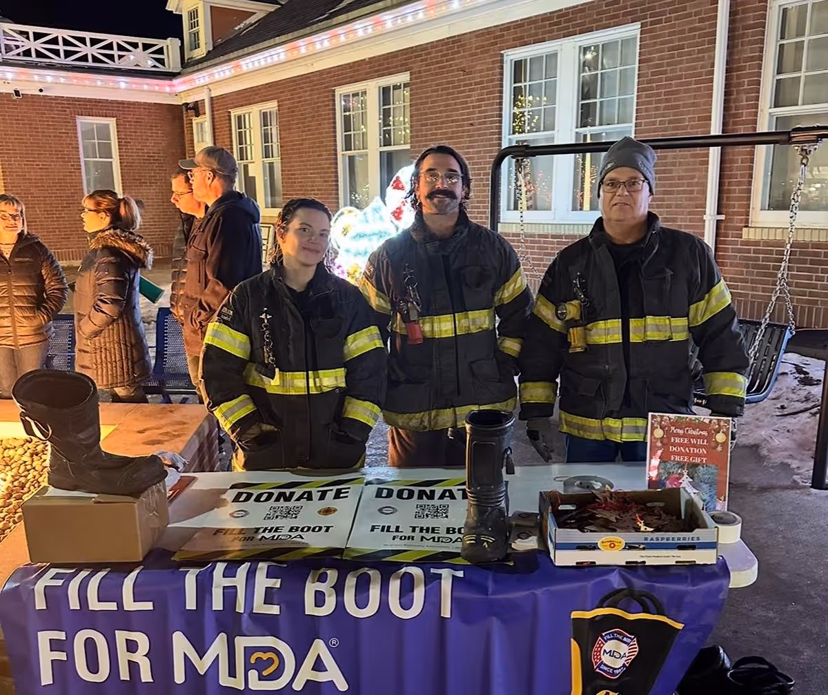 Three people in firefighter jackets standing behind a table with boots for collecting donations for MDA Fill the Boot campaign at night.
