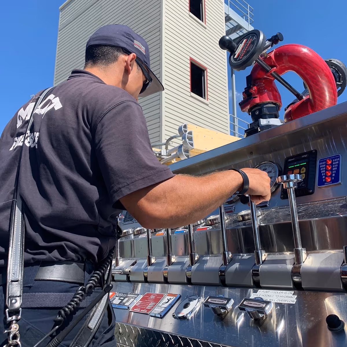 Firefighter operating controls on a fire truck pump panel outdoors near a training tower.