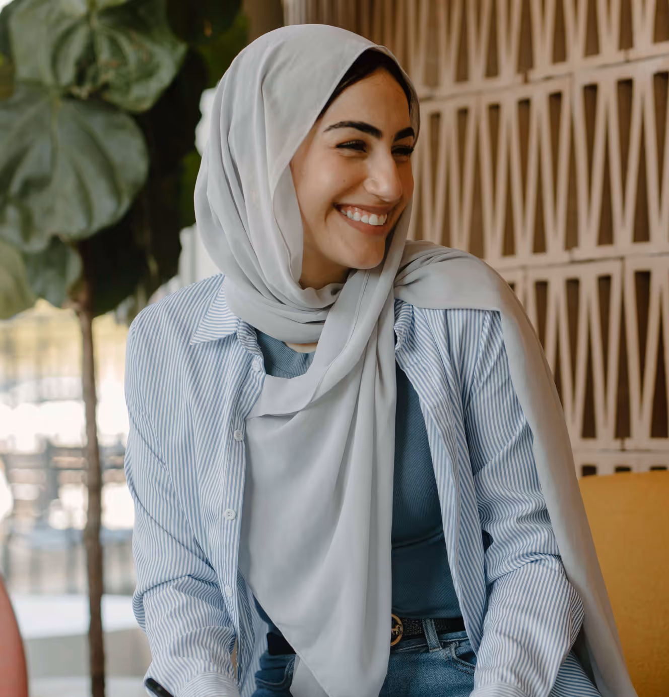 Smiling woman wearing a light gray hijab, blue striped shirt, and blue top, sitting indoors by a patterned wall and a large green plant.