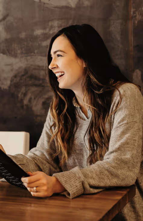 Smiling woman with long hair wearing a gray sweater sitting at a wooden table holding a black tablet.