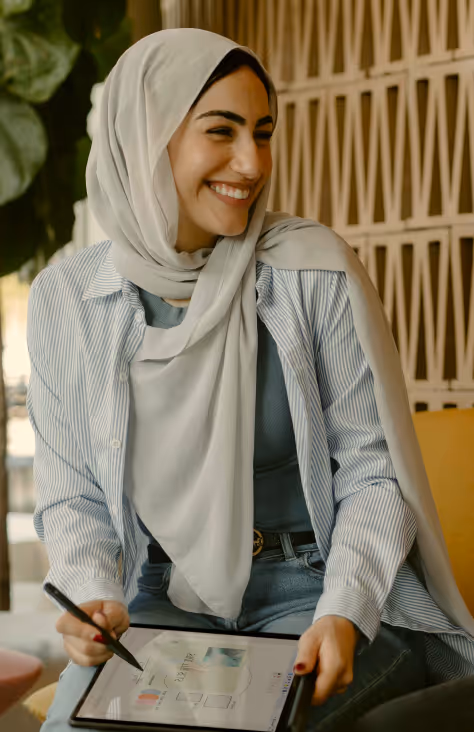 Smiling woman wearing a light-colored hijab and striped shirt using a digital tablet with a stylus pen.