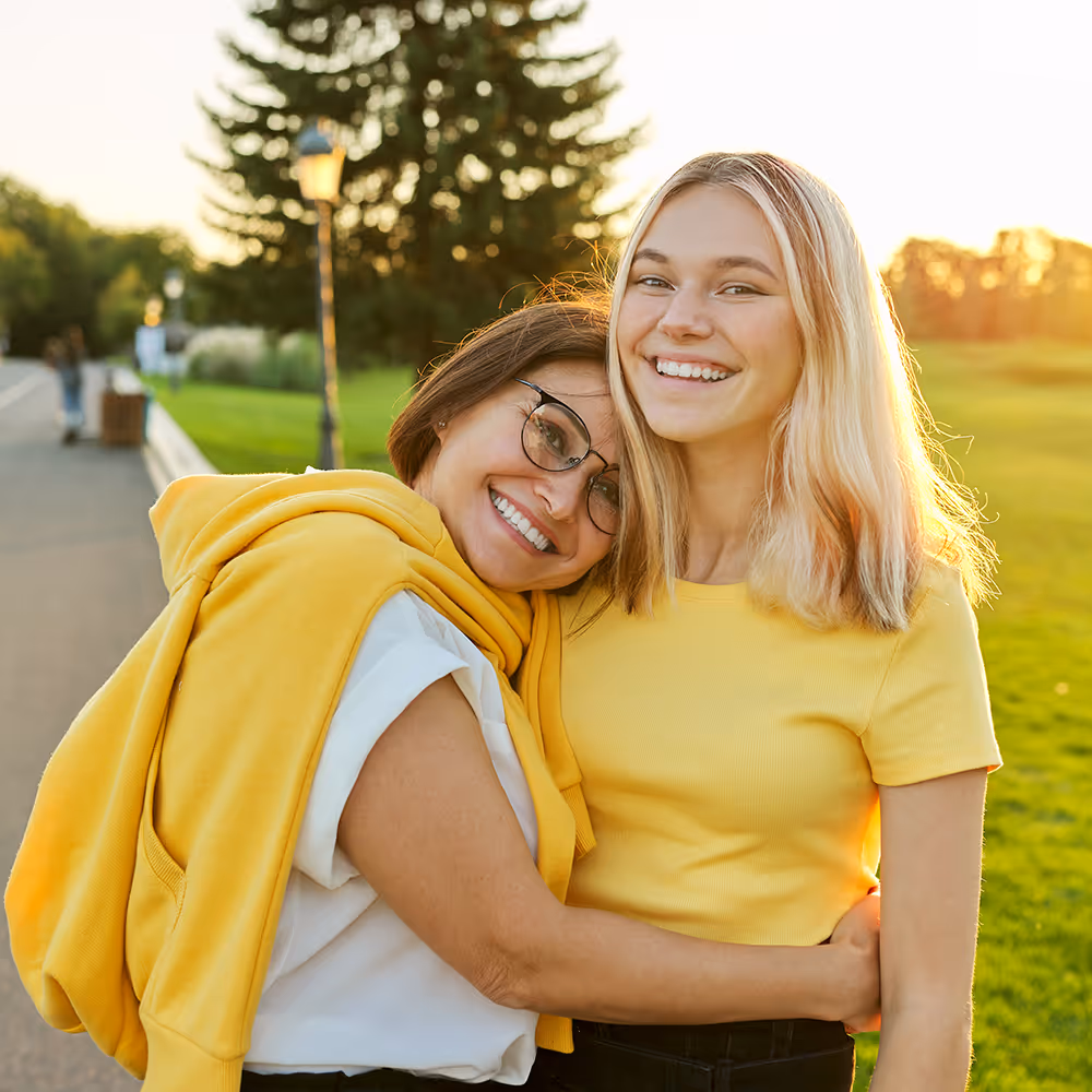 Two smiling women embracing outdoors in a park during golden hour, both wearing yellow tops.