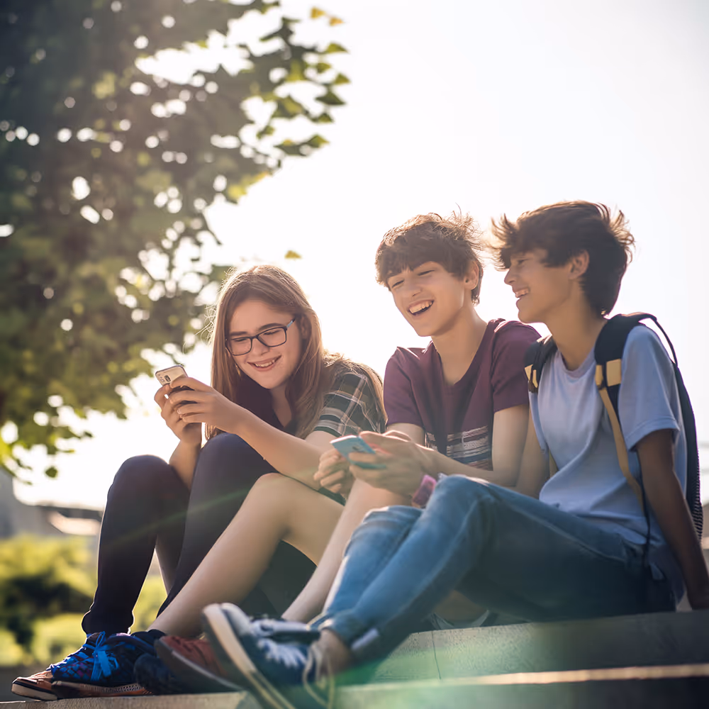 Three teenagers sitting outdoors on a ledge, smiling and looking at their smartphones on a sunny day.