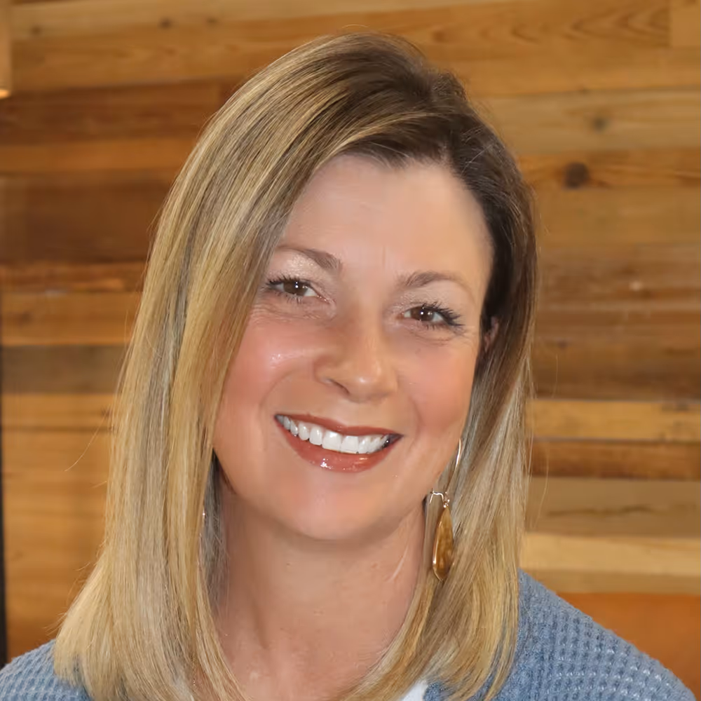 Smiling woman with shoulder-length blonde hair wearing a blue sweater and brown teardrop earrings, with a wooden background.