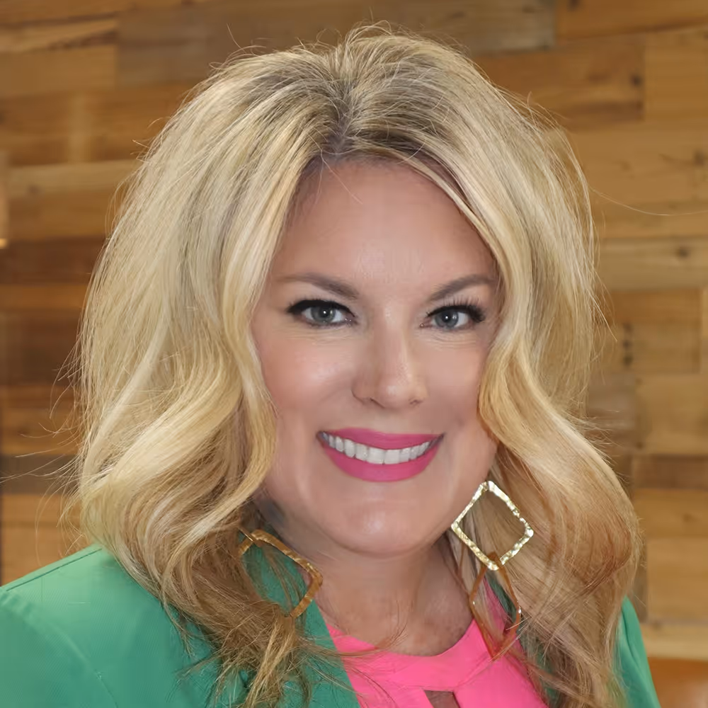 Smiling woman with blonde wavy hair, wearing bright pink lipstick, gold square earrings, a green blazer, and a pink top in front of a wooden background.
