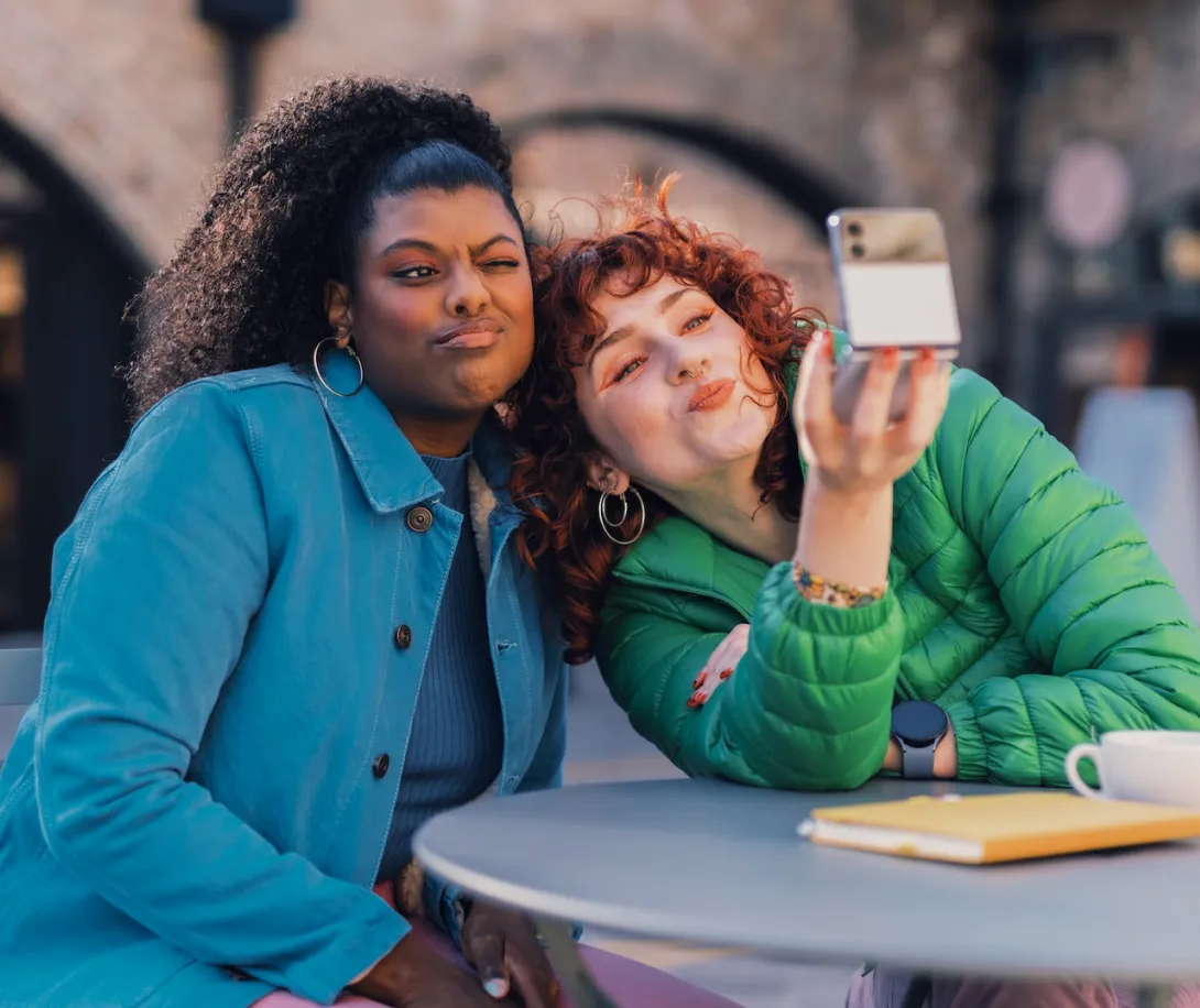 Two women sitting at an outdoor table making playful faces while taking a selfie with a smartphone.