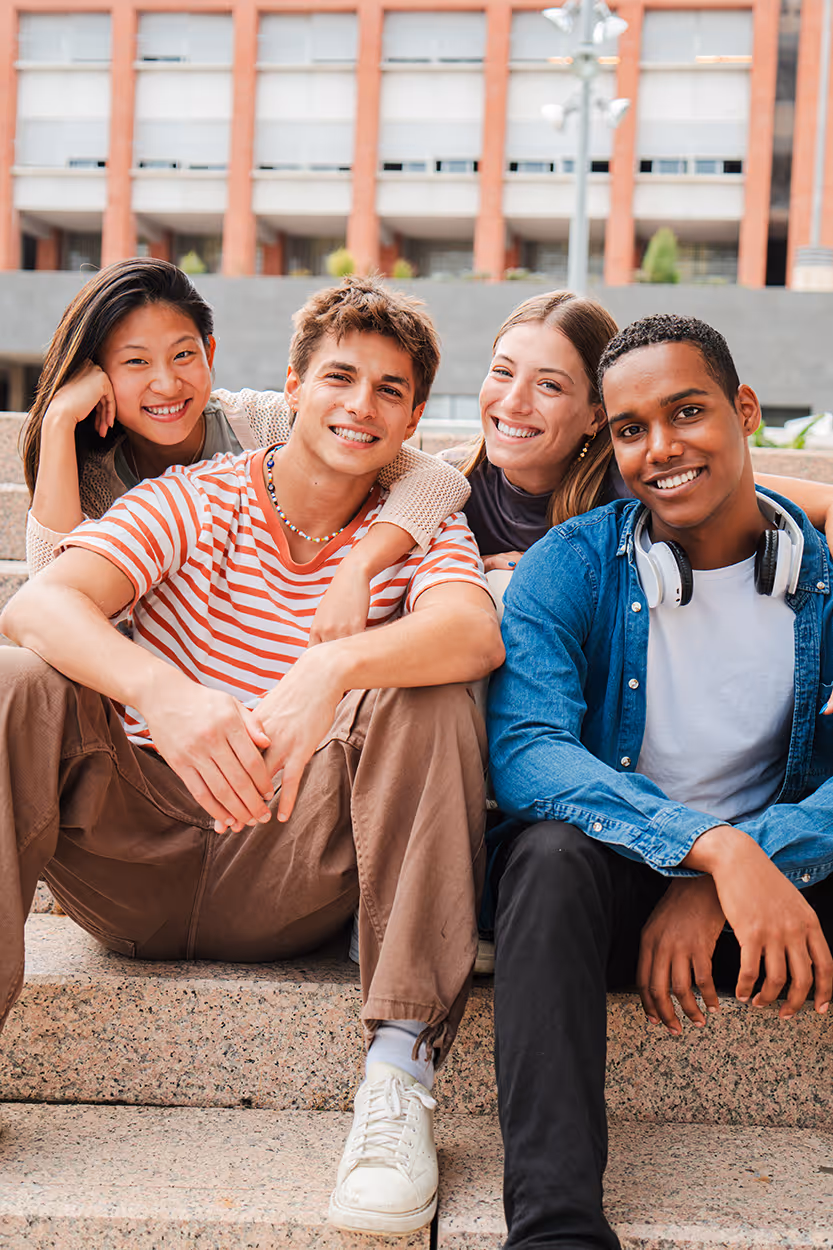 Four diverse young adults sitting on outdoor steps smiling and posing closely together.