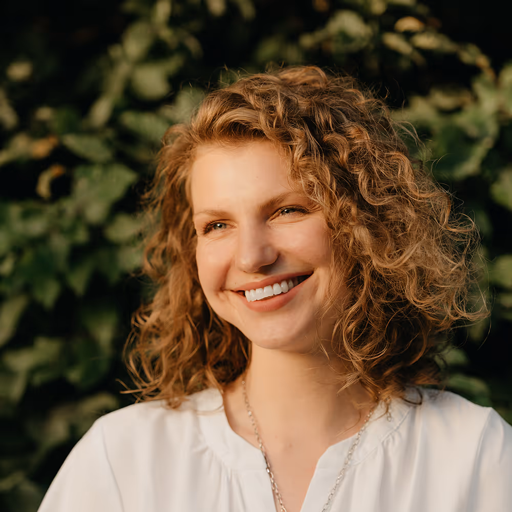 Smiling woman with curly hair wearing a white blouse and silver necklace outdoors with green foliage background.