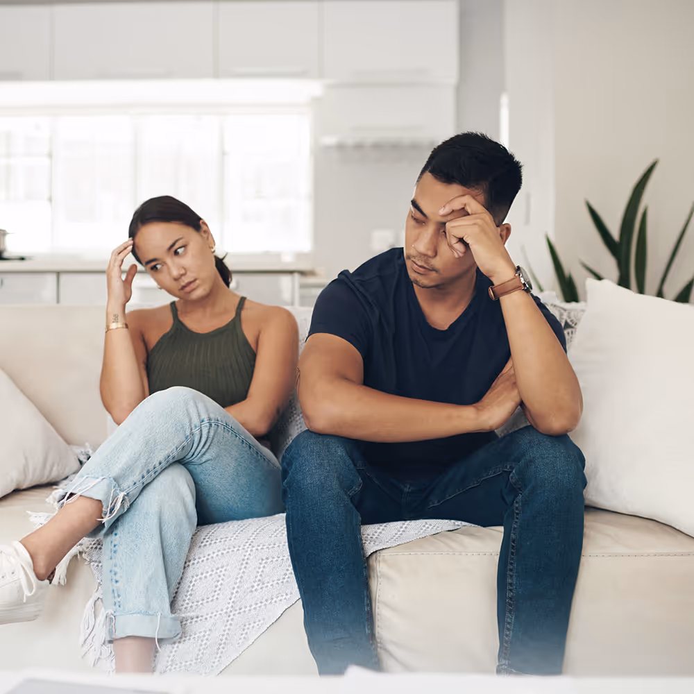 Young man and woman sitting on a couch looking upset and avoiding eye contact inside a living room.