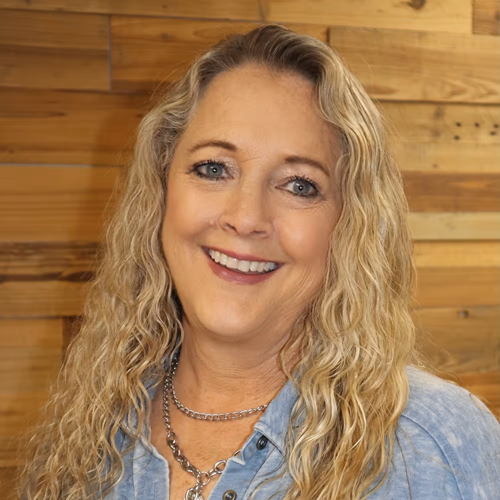 Smiling woman with long wavy blonde hair wearing a light blue denim shirt and silver chain necklaces against a wooden background.