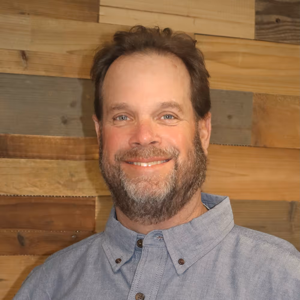 Smiling man with beard wearing a light blue button-up shirt, standing in front of a wooden plank wall.