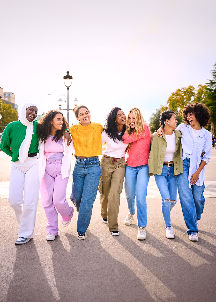 Group of six diverse young women walking arm in arm outdoors, smiling and laughing together.