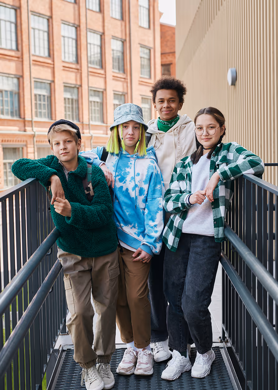 Group of four diverse teenagers standing closely on an outdoor walkway with buildings in the background, dressed in casual, colorful streetwear.