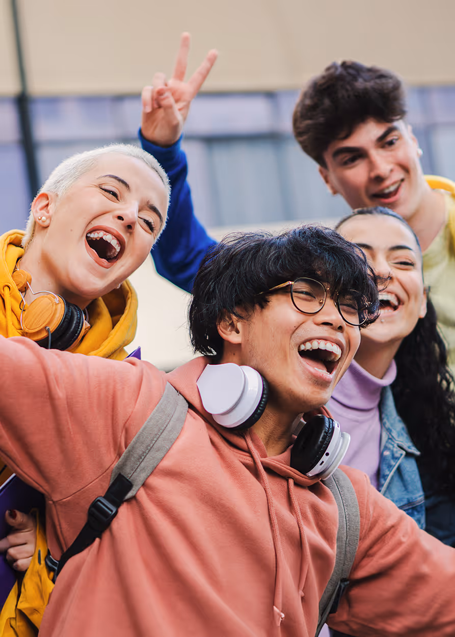 Group of four diverse young adults laughing and posing together outdoors with headphones around their necks.
