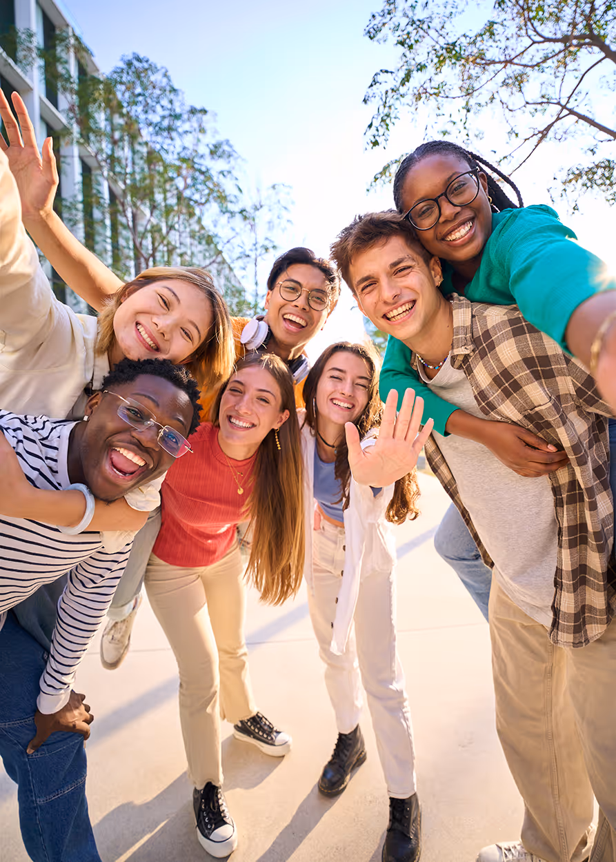 Group of seven diverse young adults smiling and waving at the camera outdoors on a sunny day.