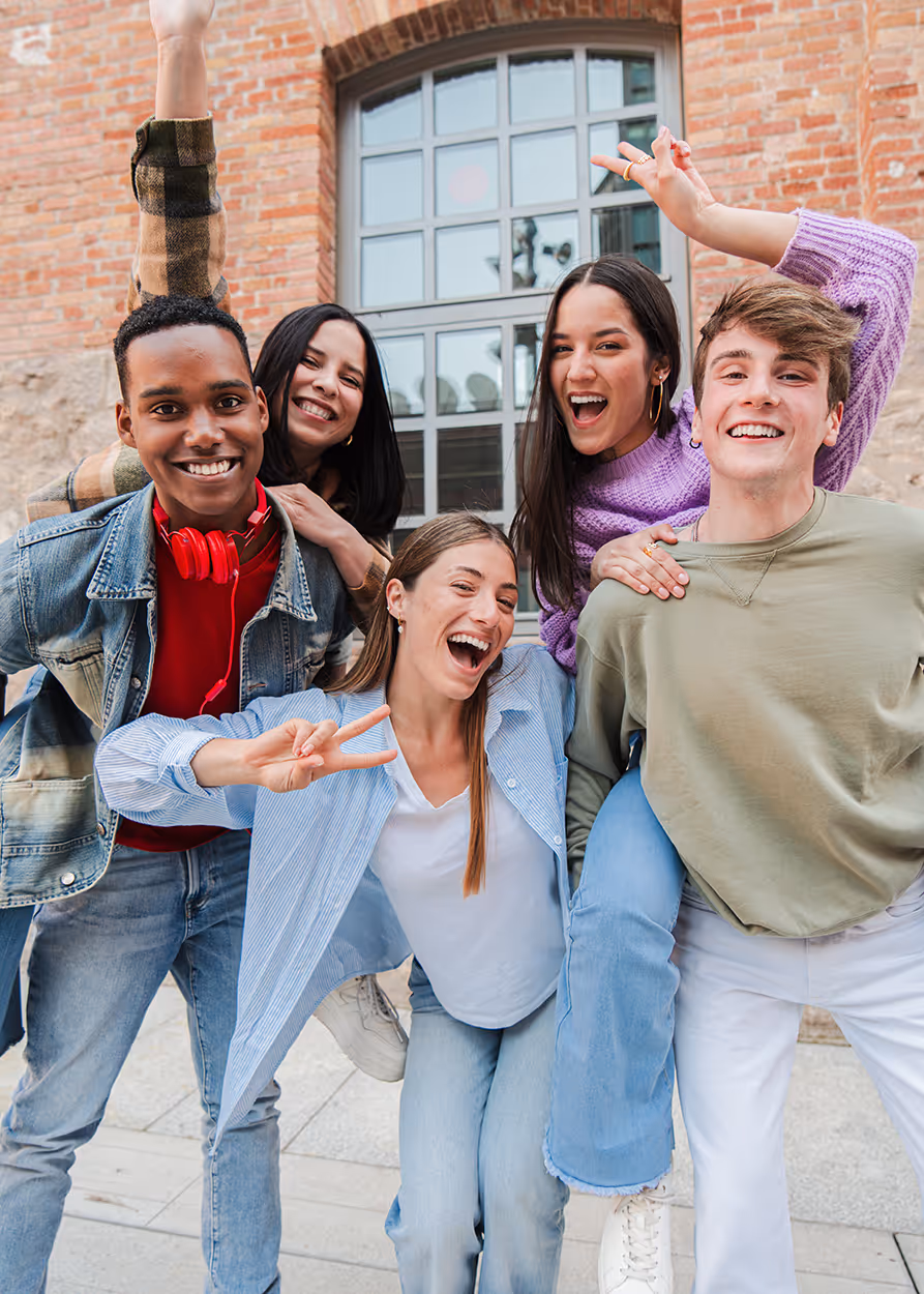 Group of five smiling young adults posing happily outdoors in front of a brick building window.