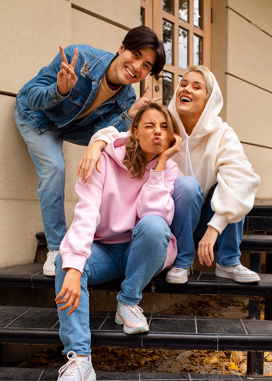 Three young adults posing playfully on outdoor stairs, with one in a pink hoodie making a funny face, another in a white hoodie smiling, and a third in a denim jacket flashing a peace sign.