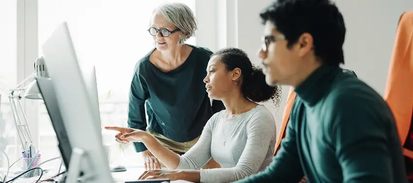 Three colleagues collaborate at a computer in a bright office, with one woman pointing at the screen.