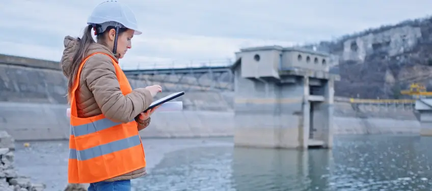 Woman in safety helmet and orange reflective vest using a tablet near a dam structure over a body of water.