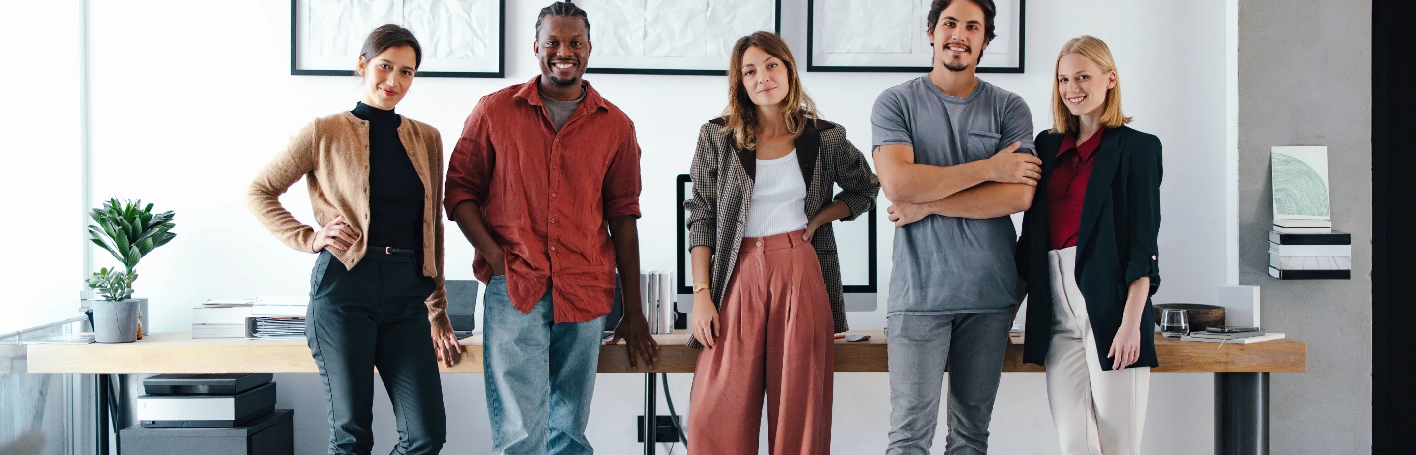 Five diverse young professionals standing confidently in a modern office with a wooden desk and computer behind them.