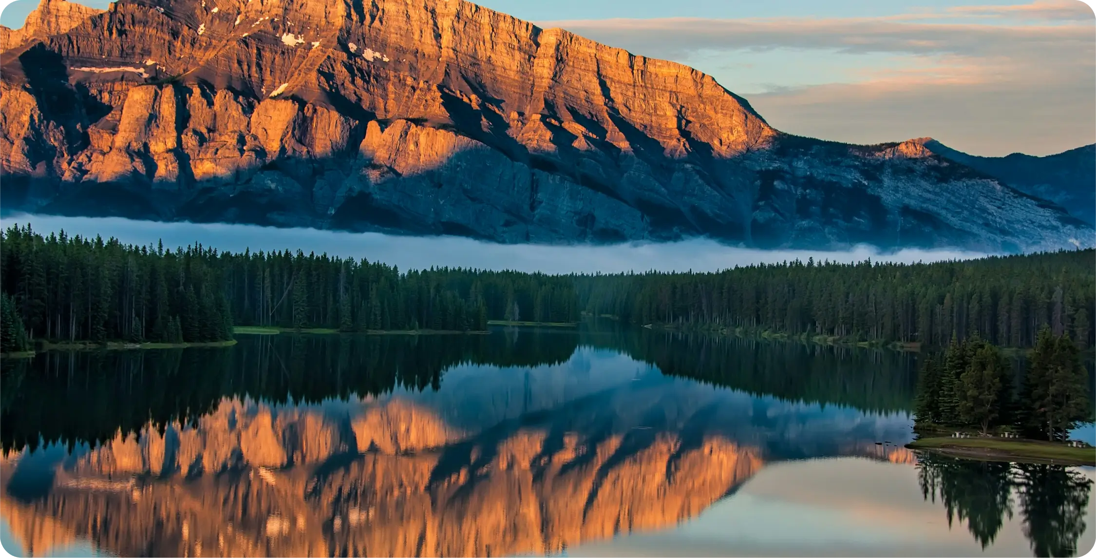 Montagne rocheuse éclairée par le soleil qui se reflète dans un lac calme avec une dense forêt de pins à sa base et une petite île bordée d'arbres sur la droite.