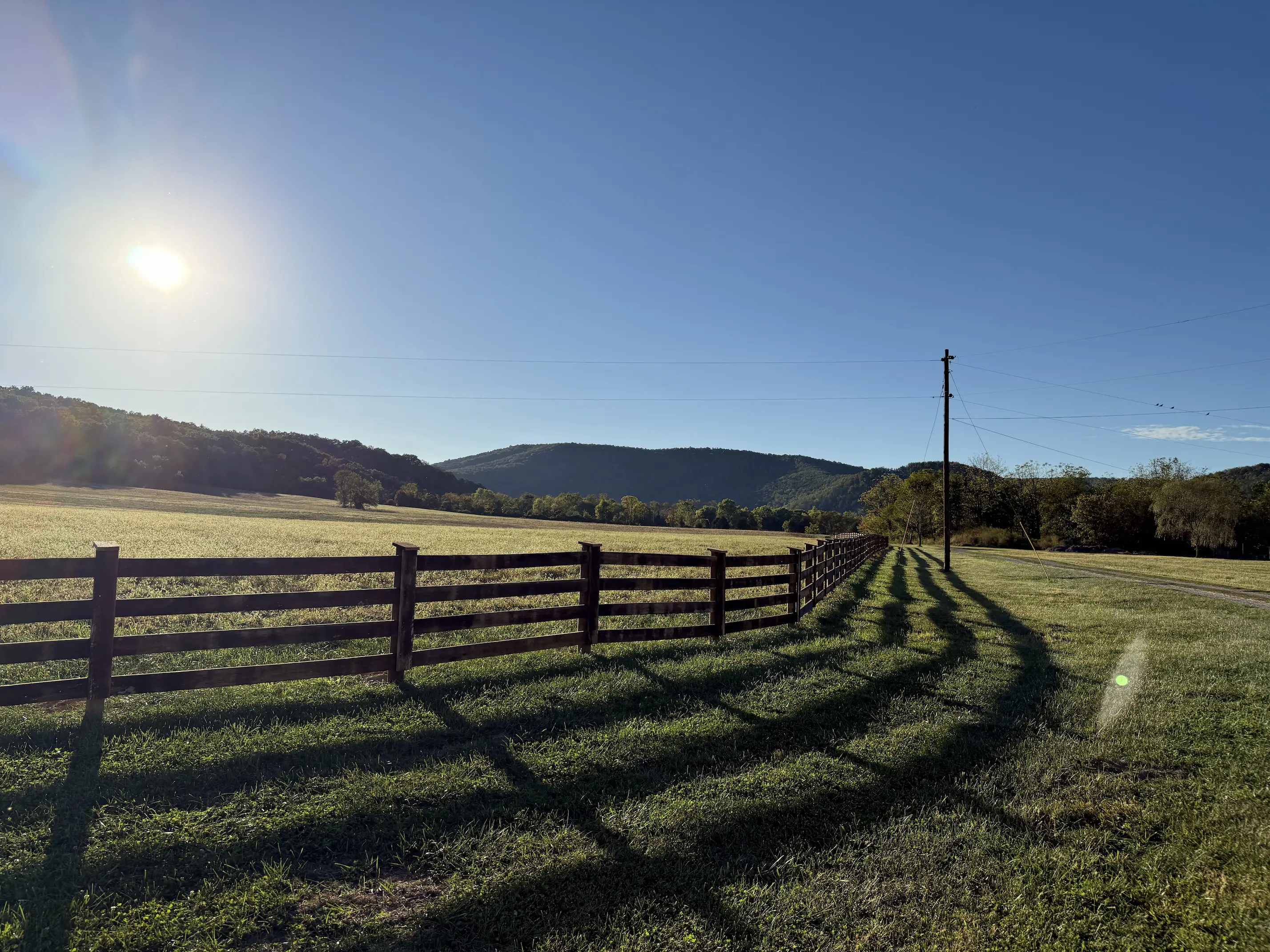 Sunlit rural field with wooden fence casting long shadows and hills in the background under clear blue sky.