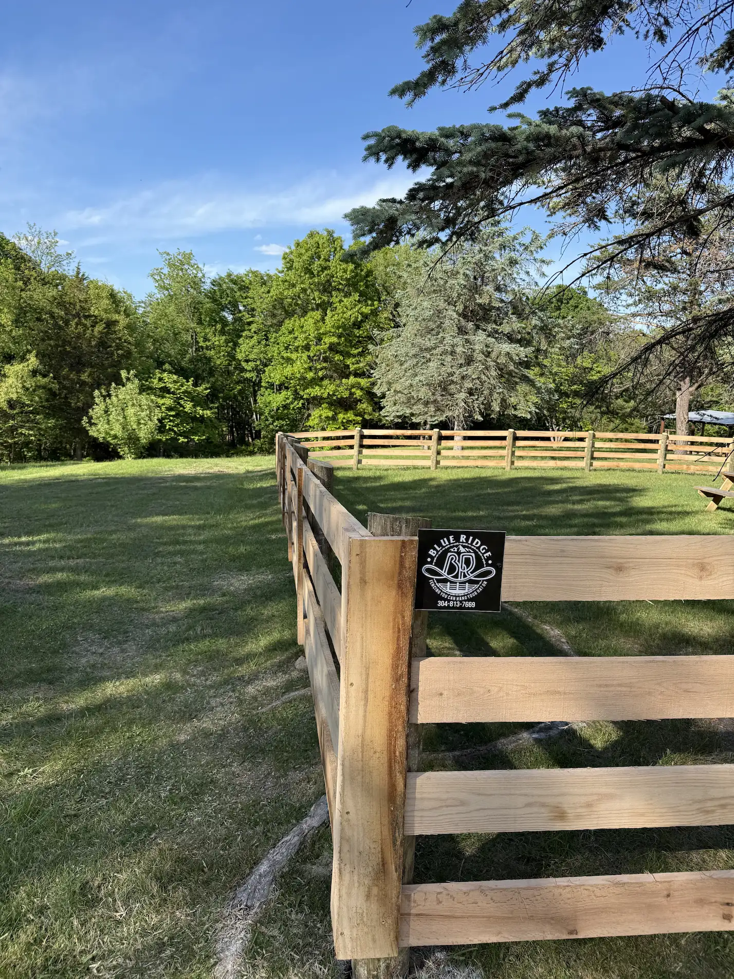 Wooden fence enclosing grassy area surrounded by green trees under blue sky.
