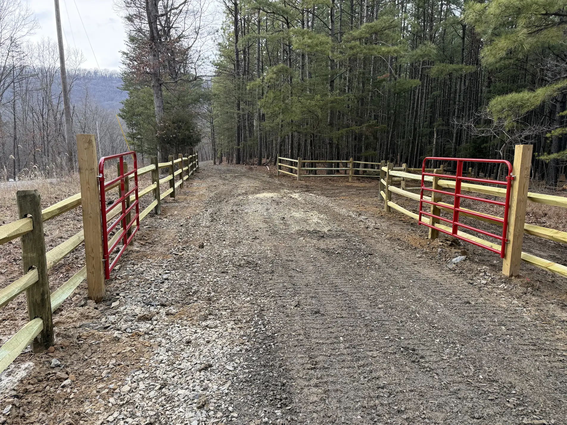 Gravel road entrance with red metal gates open between wooden fences in a forested rural area.