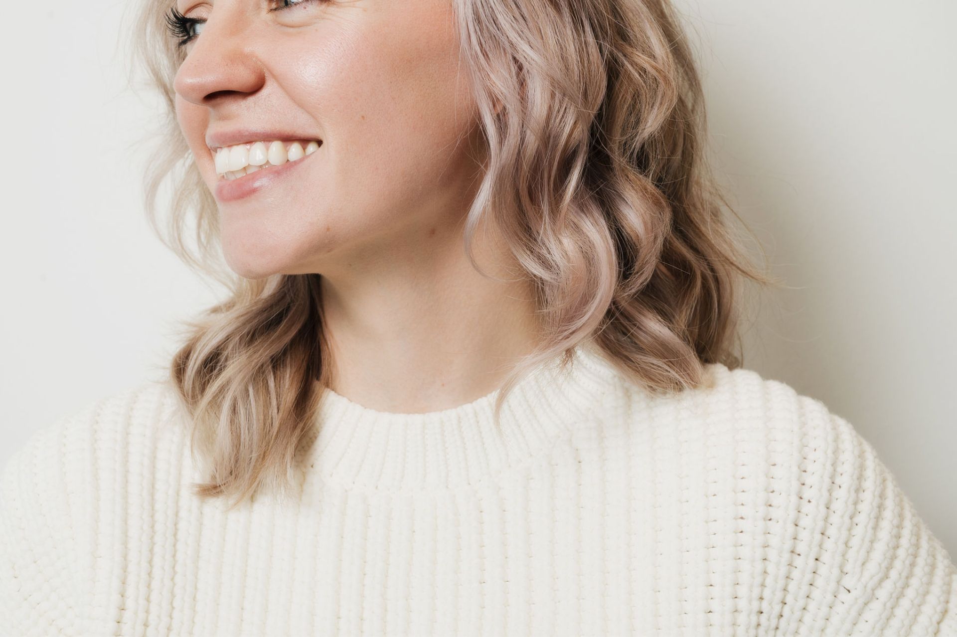Smiling woman with wavy light blonde hair wearing a white knit sweater against a light background.