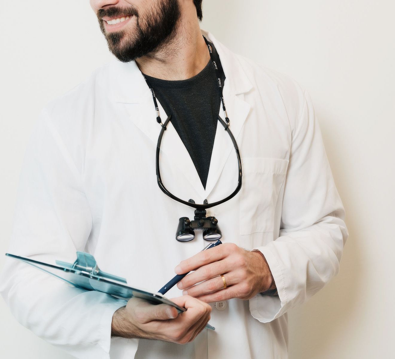 Man in a white lab coat holding a pen and a clipboard, wearing dental loupes around his neck.