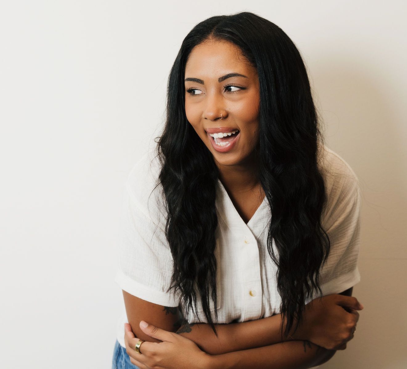 Smiling woman with long black hair wearing a white shirt, standing with arms crossed against a white wall.