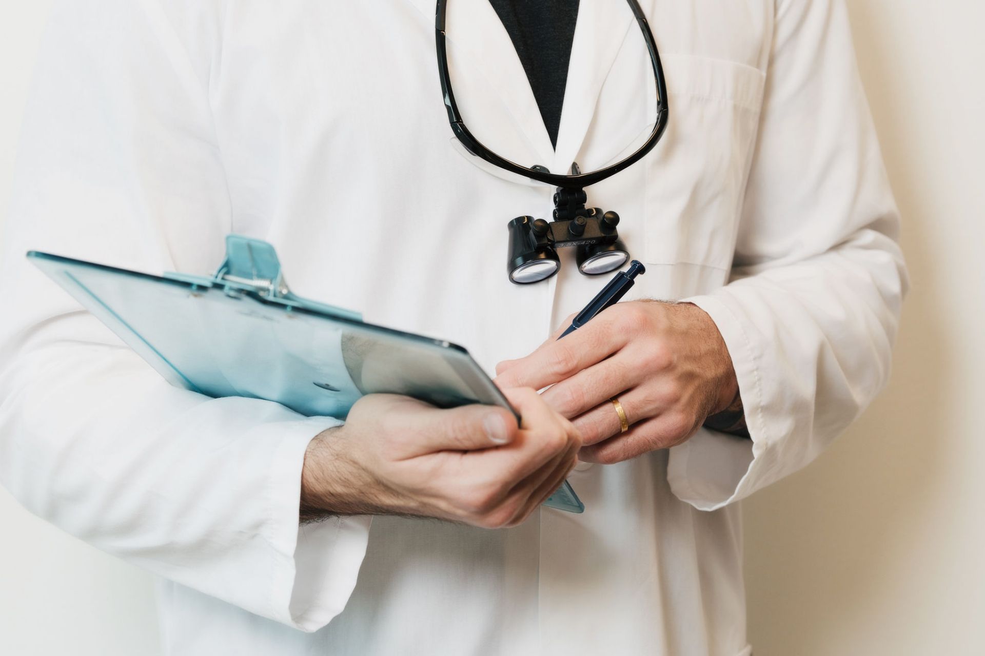 Doctor in a white coat holding a clipboard and pen, wearing magnifying glasses around the neck.