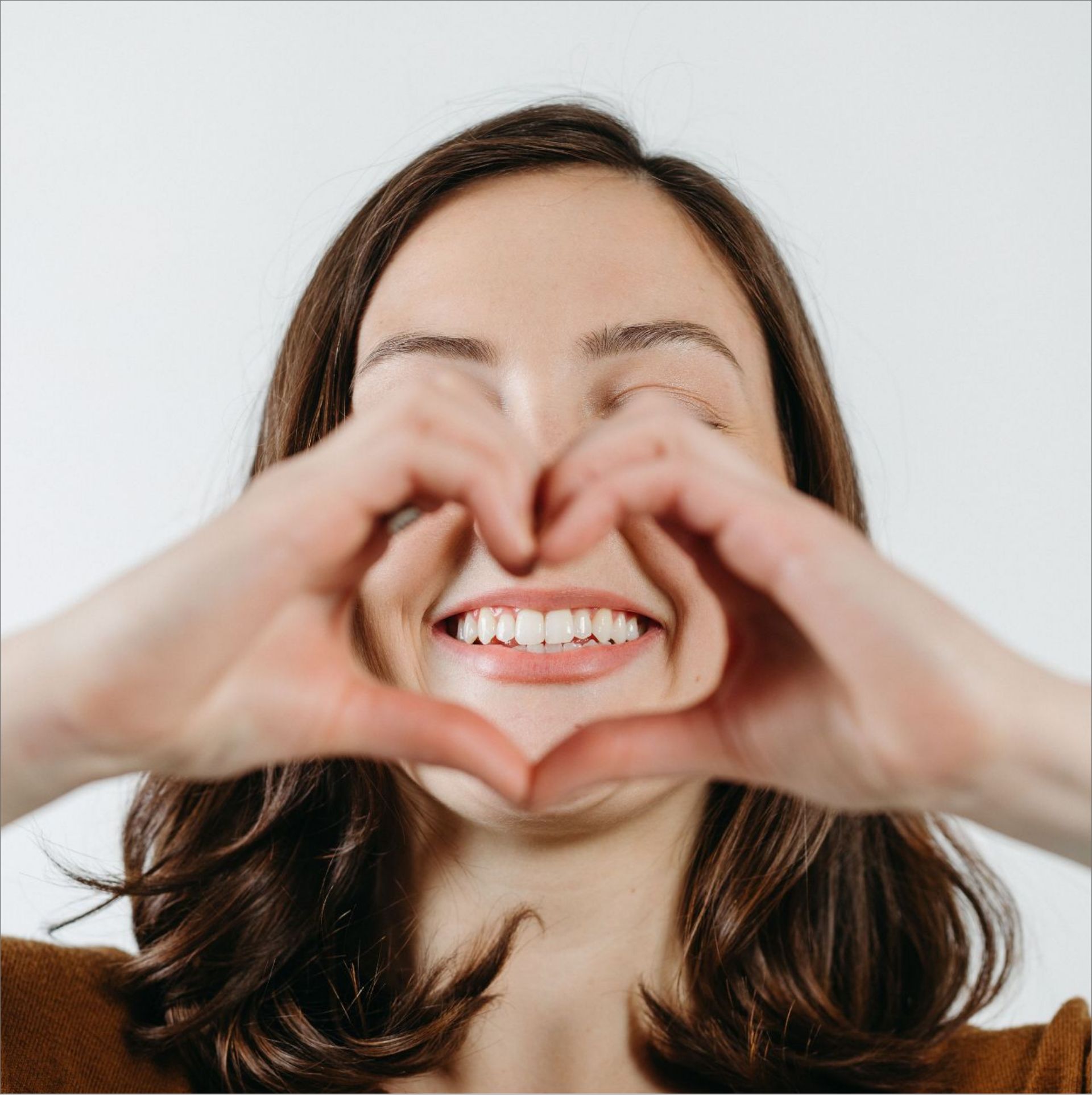 Smiling woman with brown hair forms a heart shape with her hands in front of her face.